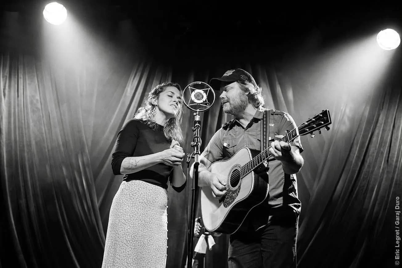 Les deux artistes de Silly Love Songs sur scène, chantant ensemble au même micro, guitare acoustique en main — photo noir et blanc
